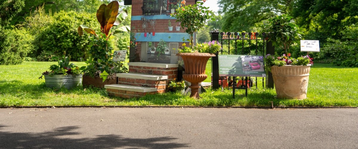 A small faux stoop is surrounded by potted plants on a lawn behind a path.