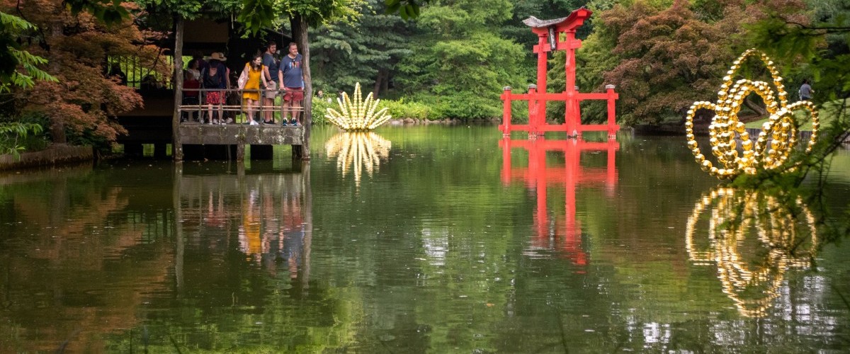 Visitors stand on a pavilion over a pond looking at two gold beaded sculptures in the form of giant lotuses reflecting in the water.