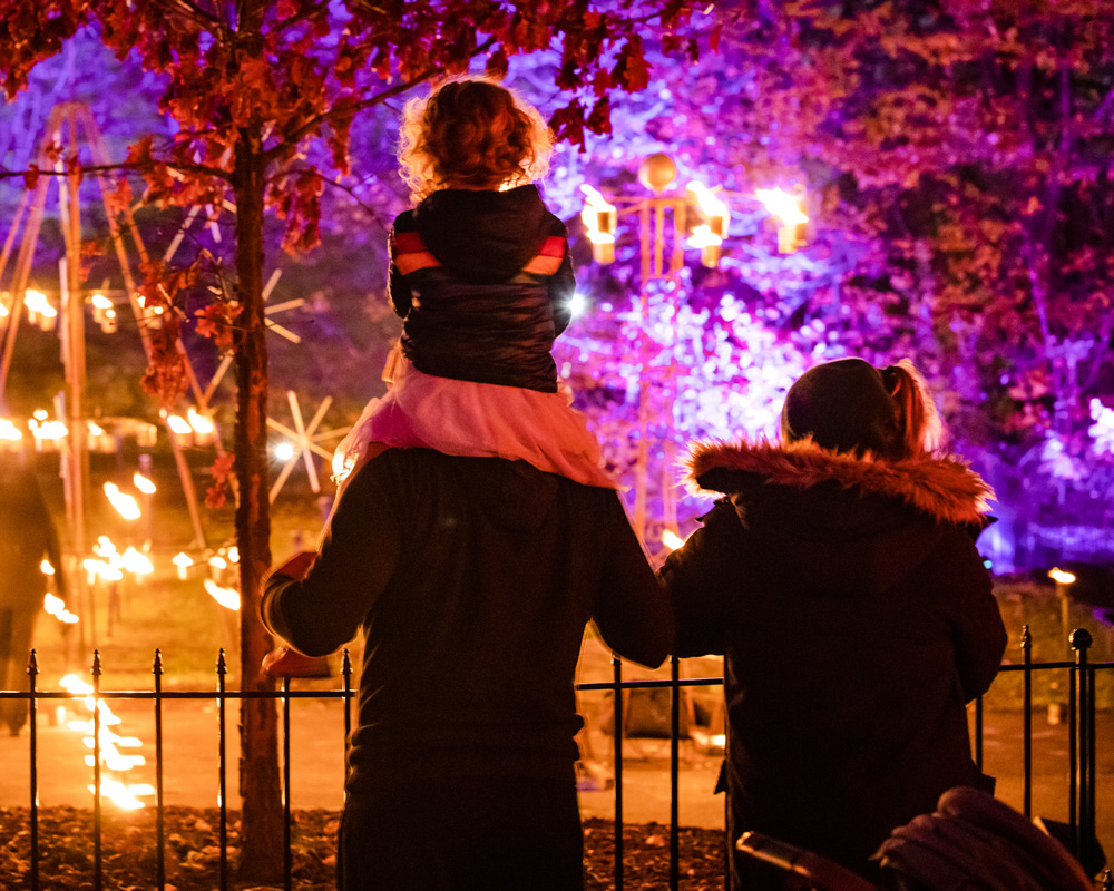 A child sits on an adult's shoulders outdoors at night while they watch mesmerizing pots of flame.