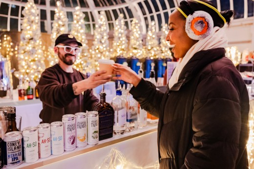 A woman in a winter coat takes a drink from a bartender in a room full of twinkling white lights